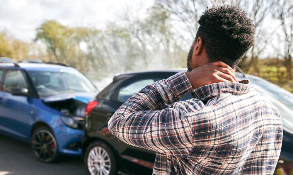 Young man rubbing neck in pain from whiplash injury standing by damaged car after traffic accident